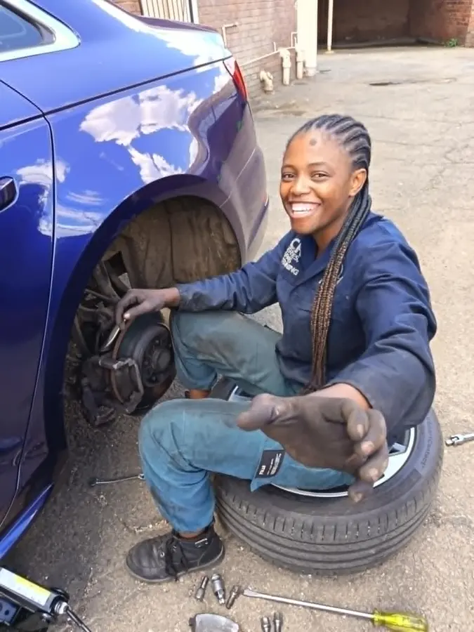 A light vehicle mechanic crouched beside a blue car removing a wheel, smiling at the camera